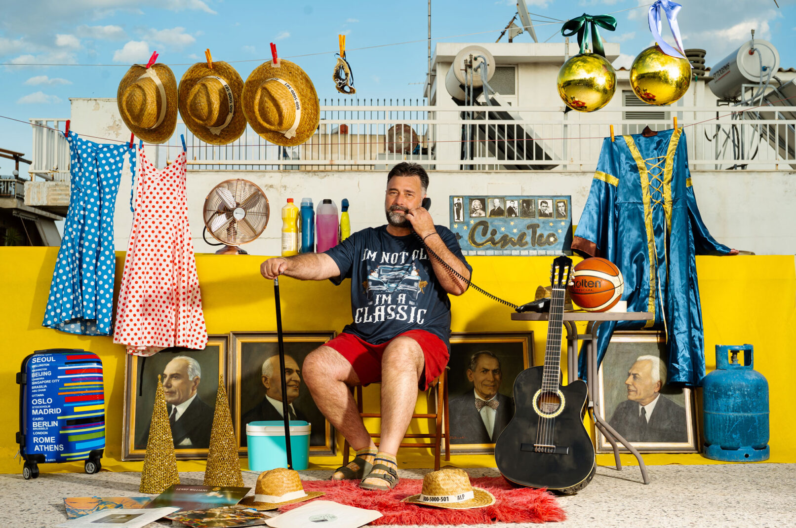 A person sitting on a terrace speaking on an old phone with many things around like paintings, guitar, hats dresses and ore